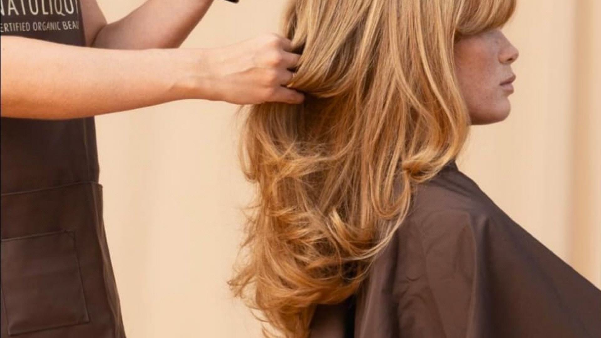 Dame mit langem Haar beim Frisör und dieser mit Spraydose hinter ihr.